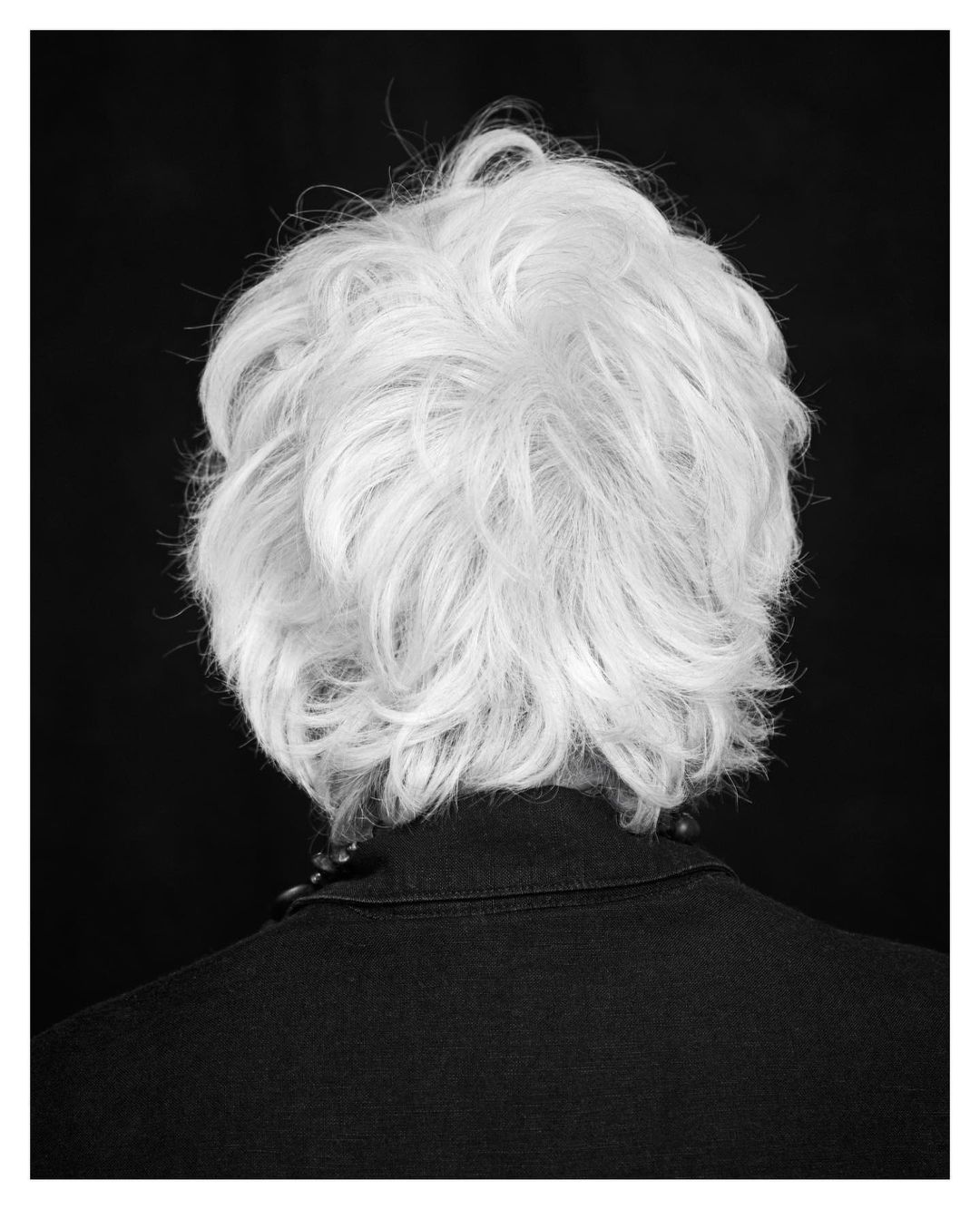 Back view of a person with short, wavy, light gray hair, wearing a dark collared shirt, against a solid black background in a studio shot.