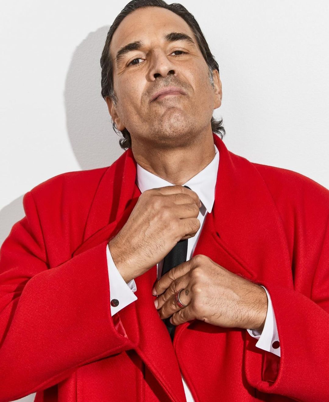 Portrait of an adult male in a red overcoat and a white shirt, adjusting a black tie against a plain white backdrop in a photo studio.