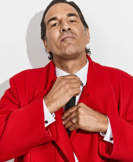 Portrait of an adult male in a red overcoat and a white shirt, adjusting a black tie against a plain white backdrop in a photo studio.