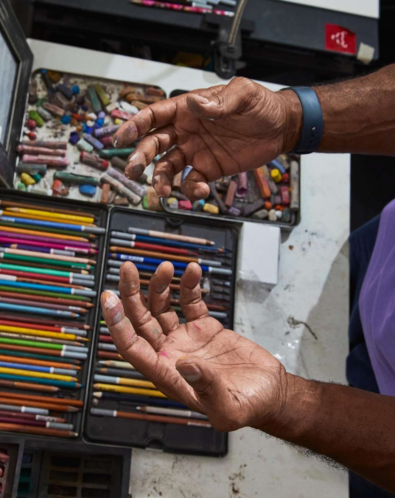Close-up of an artist's hands covered in pastel dust, positioned over a set of colorful pastels and colored pencils in metal cases.
