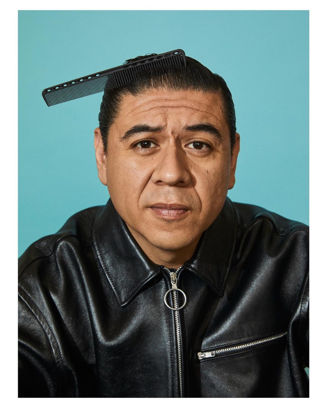Man in black leather jacket poses with comb on head against teal backdrop in studio portrait. Edgy fashion and grooming.