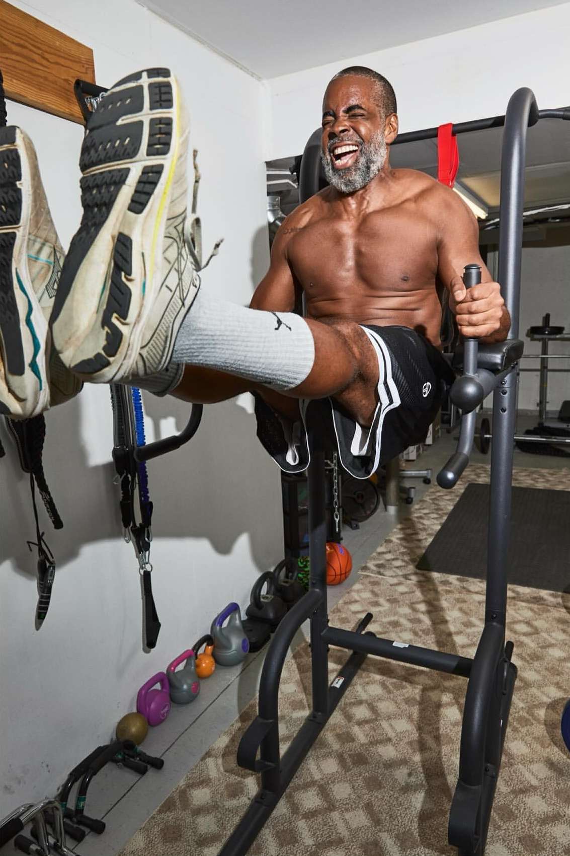 An adult man with graying beard and no shirt works out indoors on exercise equipment lifting his knees up to his chest.