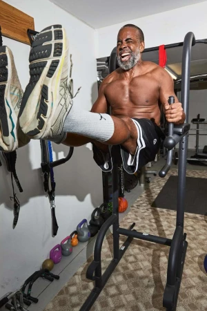 An adult man with graying beard and no shirt works out indoors on exercise equipment lifting his knees up to his chest.