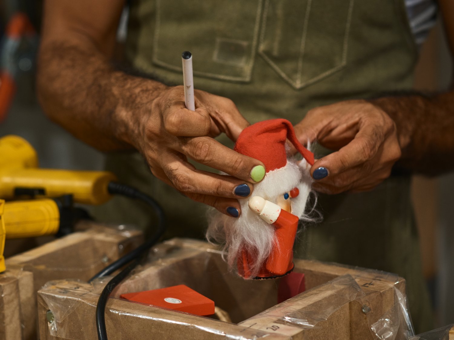 Person holds a handmade Santa Claus doll with red hat and white beard; yellow glue gun visible, indoors. Close-up shot, craft setting.