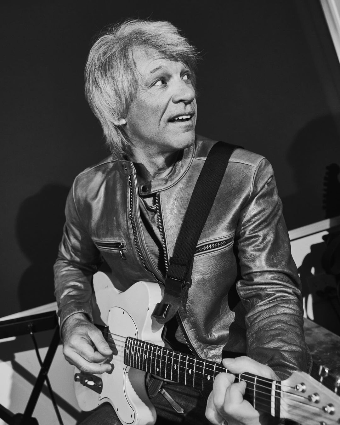Black and white portrait of Jon Bon Jovi playing a white electric guitar in a leather jacket, indoors, with a dark background.
