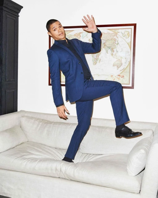 A man in a blue suit strikes a pose on a white sofa in front of a map. The photo is taken indoors with soft lighting.