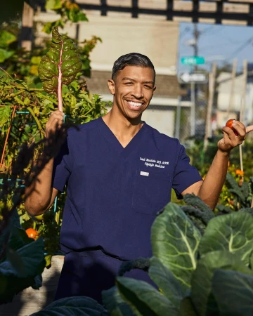 Smiling man in scrubs in a garden, holding fresh produce in sunlight. Focus on health, nutrition, and wellness in an outdoor setting.