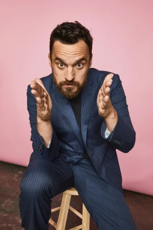 Bearded adult man in a blue pinstripe suit, sitting on a wooden stool, gesturing with his hands, set against a pink background.