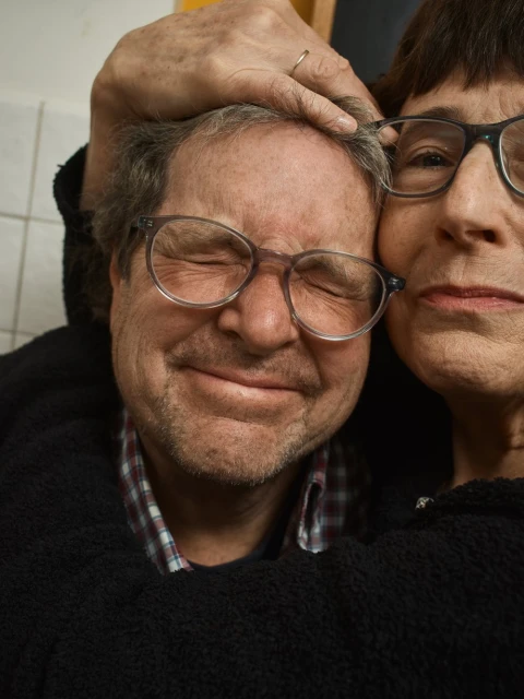Close-up of an older couple embracing indoors, both wearing glasses. The man has his eyes closed and the woman looks toward the camera.