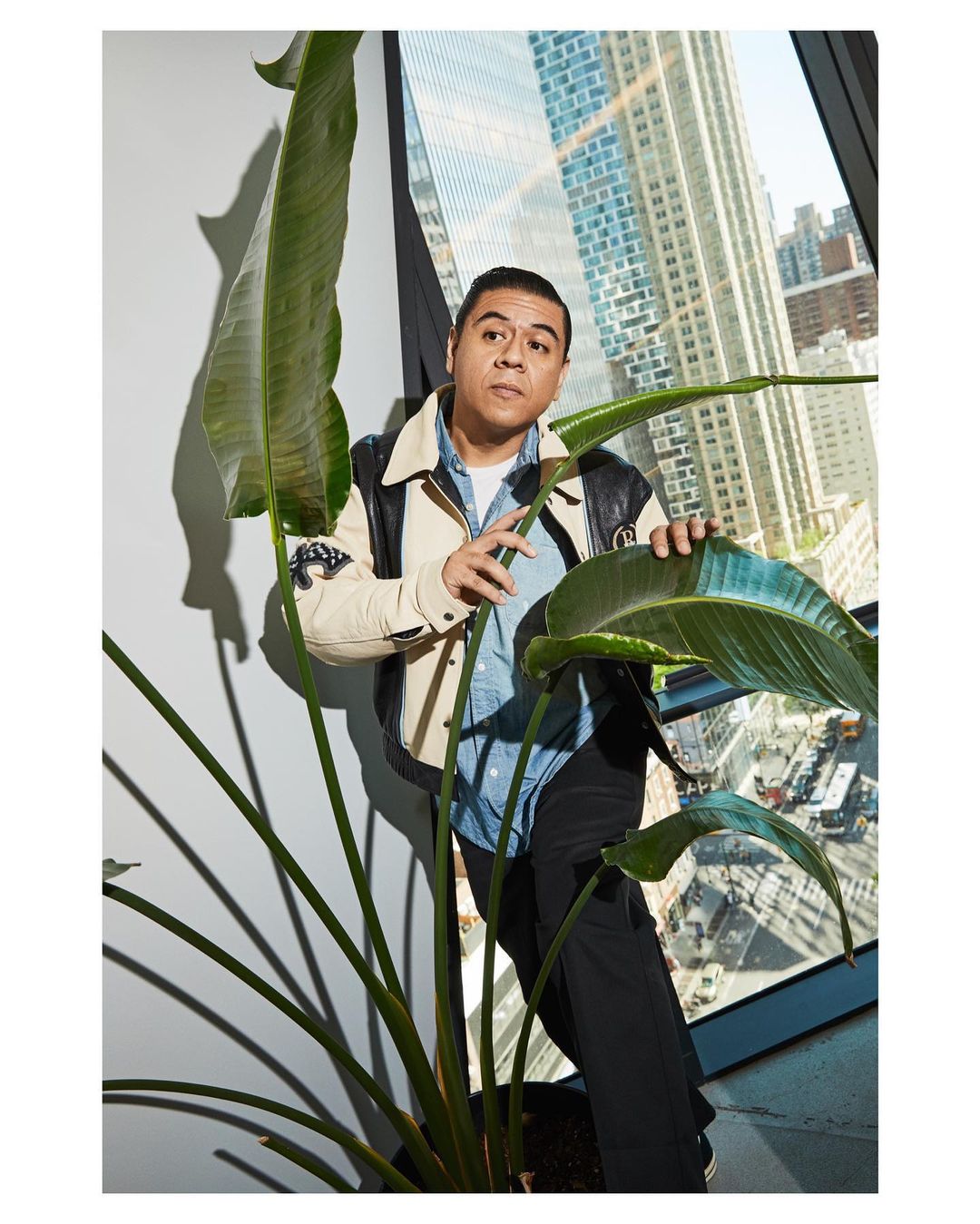 Stylish man poses indoors next to large bird of paradise plant with skyscraper visible through the window in the background.