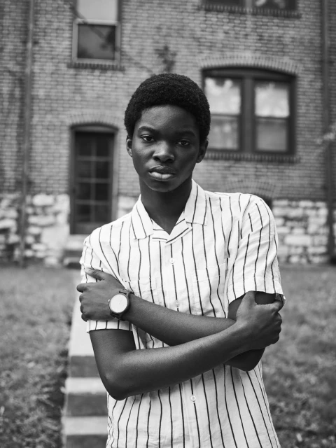 Portrait of a young Black adult with arms crossed wearing a vertically striped shirt outdoors in front of a brick building and greenery.