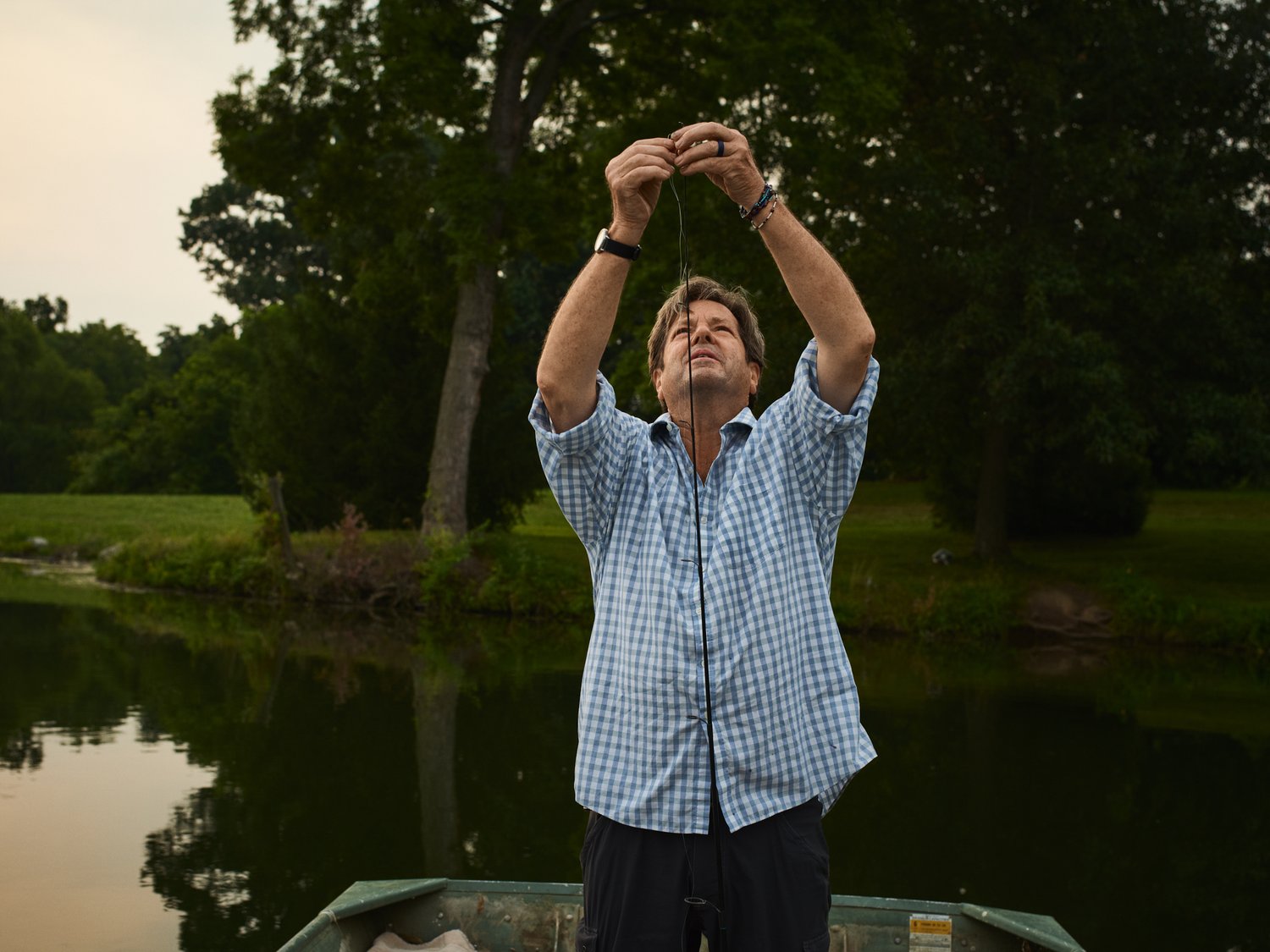 Man standing in a boat, arms raised, holding a fishing line against a backdrop of a tranquil lake and lush green trees.
