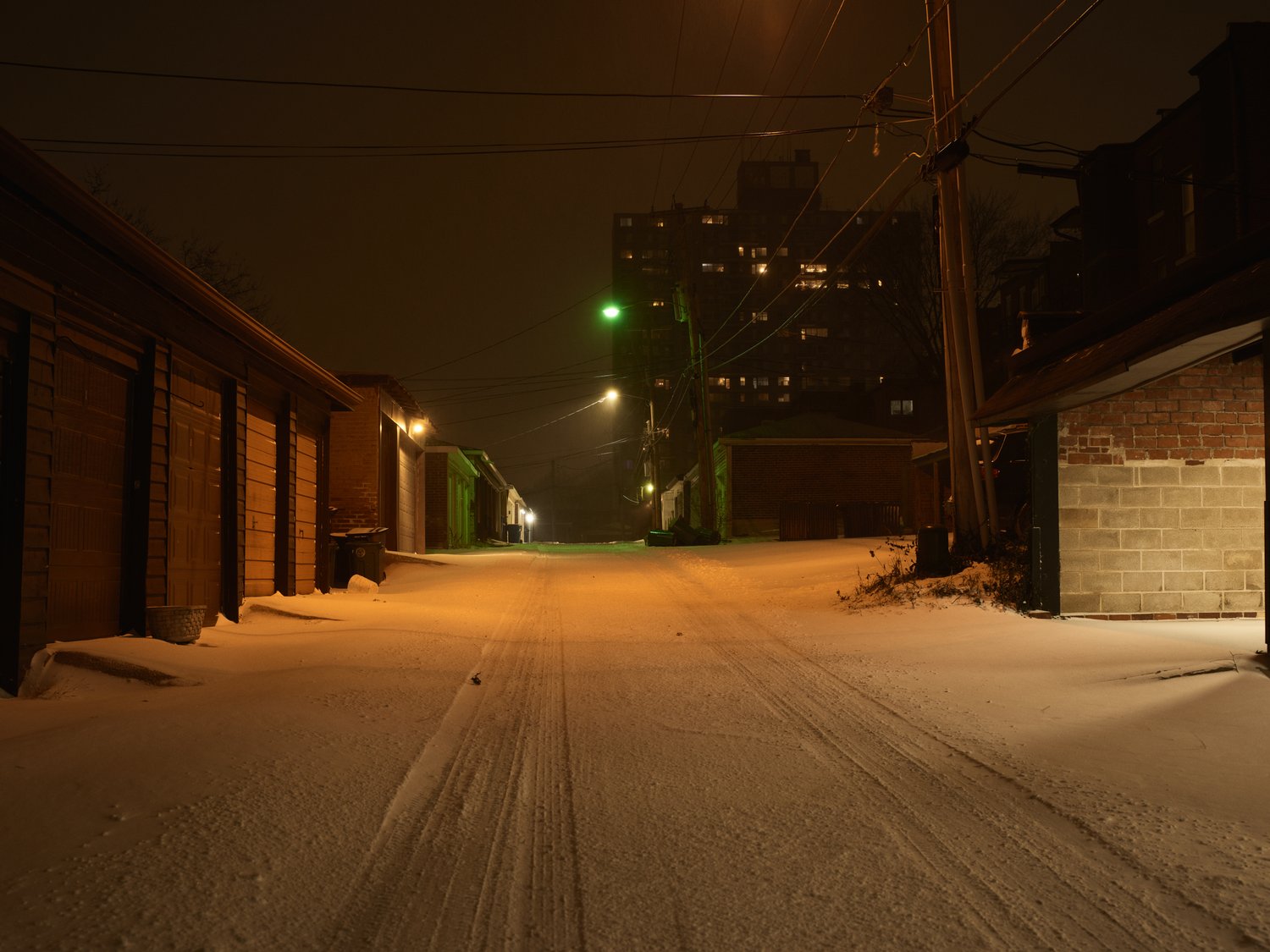 Winter night scene in an urban alleyway with snow-covered ground, flanked by garage doors and an apartment building in the distance.