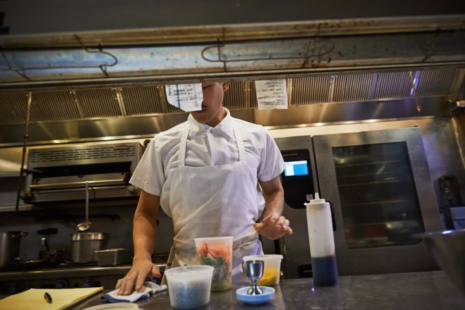 Chef in a commercial kitchen preparing food with various containers and kitchen equipment. The environment is stainless steel.