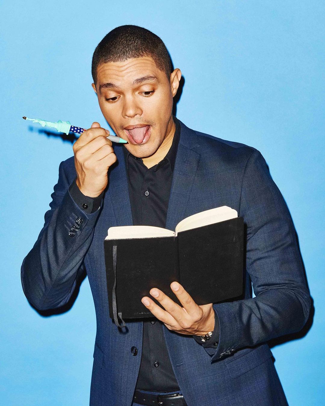Trevor Noah in a blazer and black shirt holding a black book with a pen in his mouth against a light blue studio backdrop.