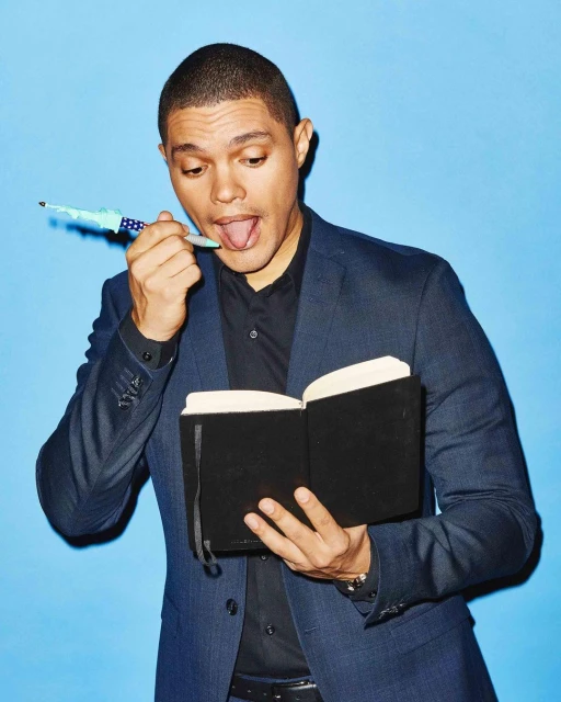 Trevor Noah in a blazer and black shirt holding a black book with a pen in his mouth against a light blue studio backdrop.