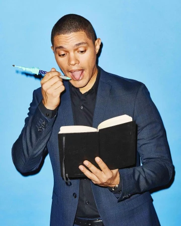 Trevor Noah in a blazer and black shirt holding a black book with a pen in his mouth against a light blue studio backdrop.