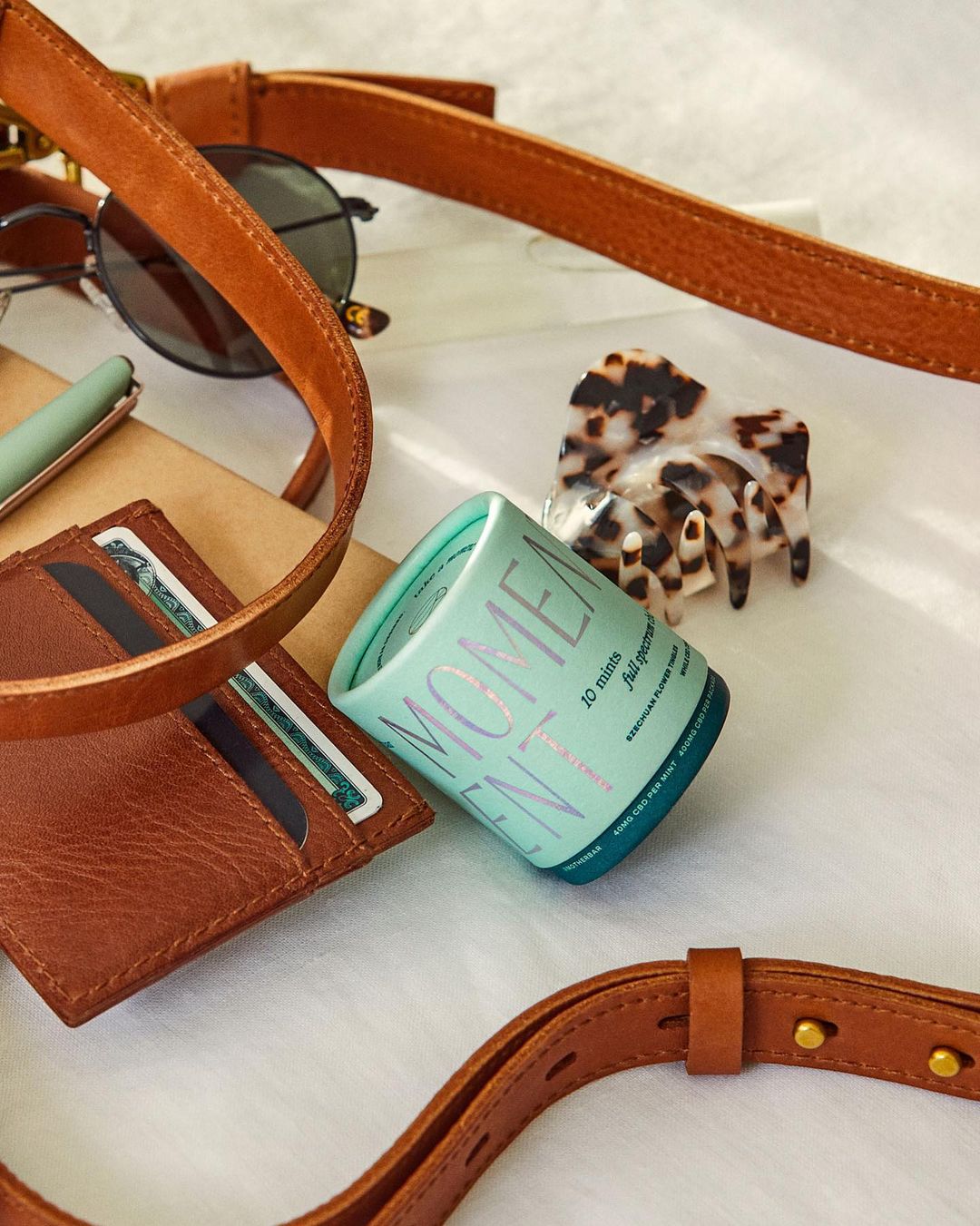 Elevated view of a brown leather wallet, belt, sunglasses, pen, hair clip, and mints on a white surface, showcasing personal accessories.