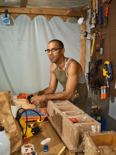 Portrait of an adult male wearing glasses and an apron, working at a wooden table with tools and craft supplies in a cluttered workshop.