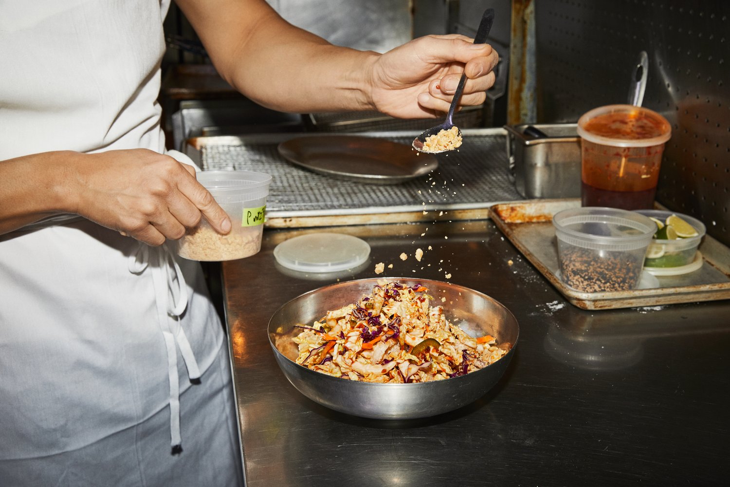 A person in a white apron adds toppings to a colorful salad in a silver bowl on a stainless steel kitchen surface.