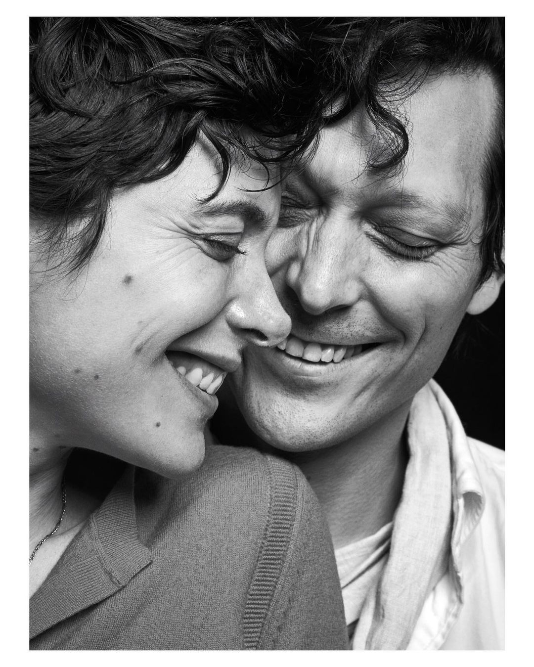 Black and white portrait of a smiling couple, a woman and a man, embracing closely with dark curly hair in an indoor setting.