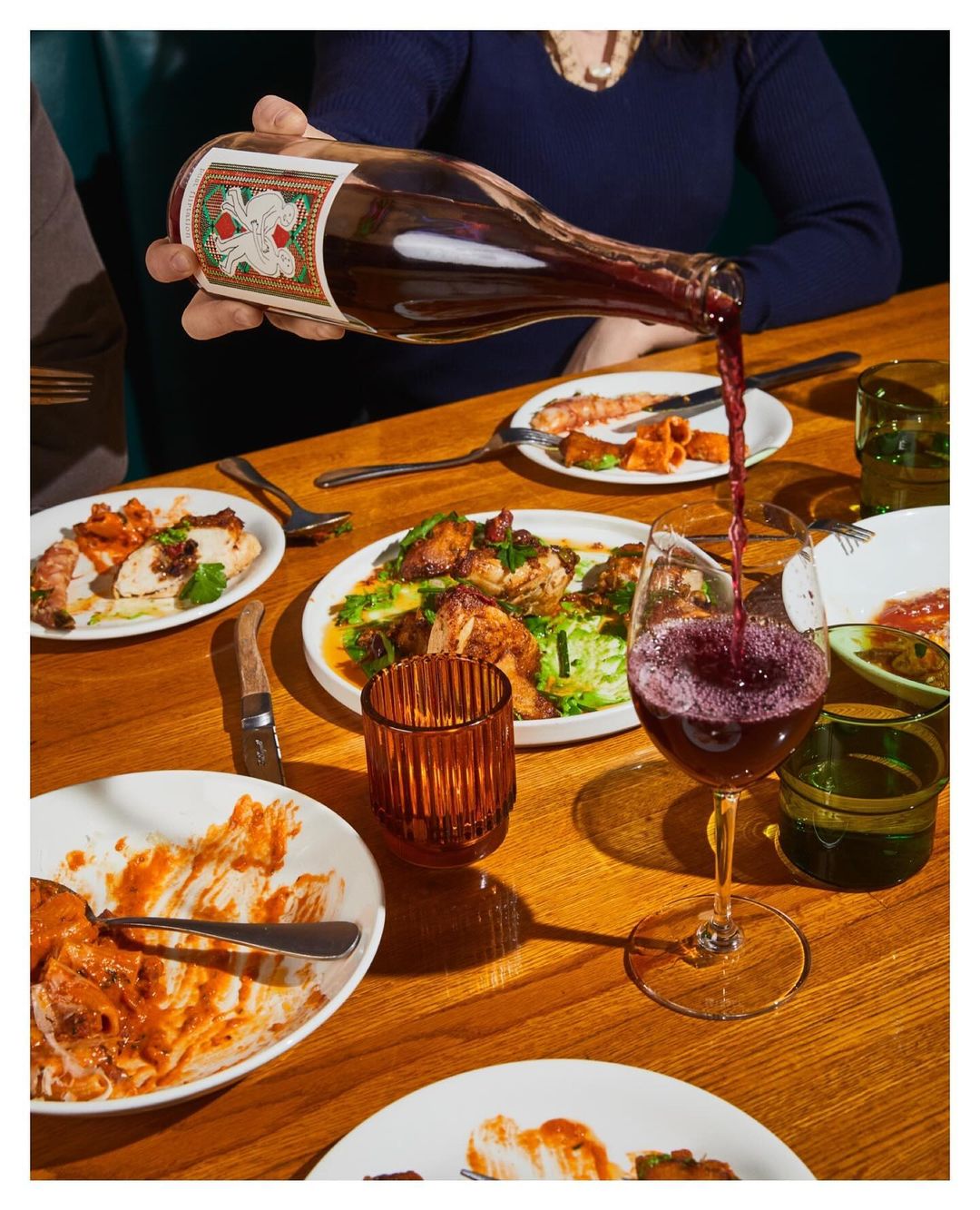 Person pouring red wine into a wine glass at a wooden table with several plates of food and glassware in a warm, indoor setting.