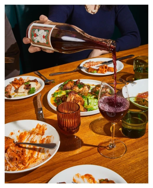 Person pouring red wine into a wine glass at a wooden table with several plates of food and glassware in a warm, indoor setting.