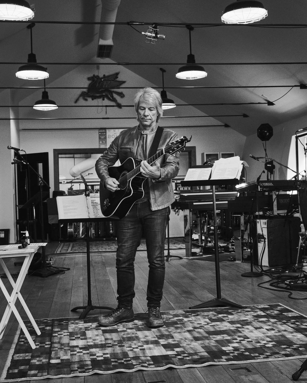 Monochrome shot of a musician playing an acoustic guitar in a music studio, illuminated by overhead lights, wearing a leather jacket and jeans.