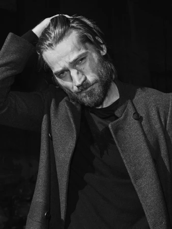 Monochrome portrait of a bearded man wearing a coat, running his hand through his hair in a dimly lit outdoor setting.