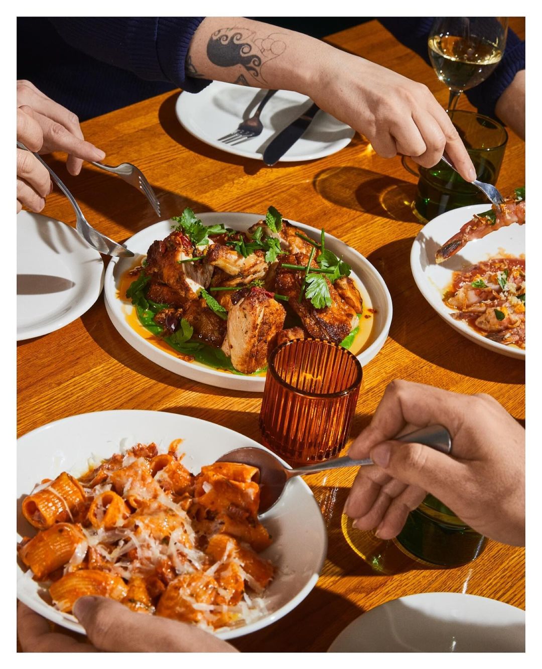 Overhead shot of a wooden table laden with plates of pasta, roasted chicken, and shrimp; shared by multiple people using silverware.