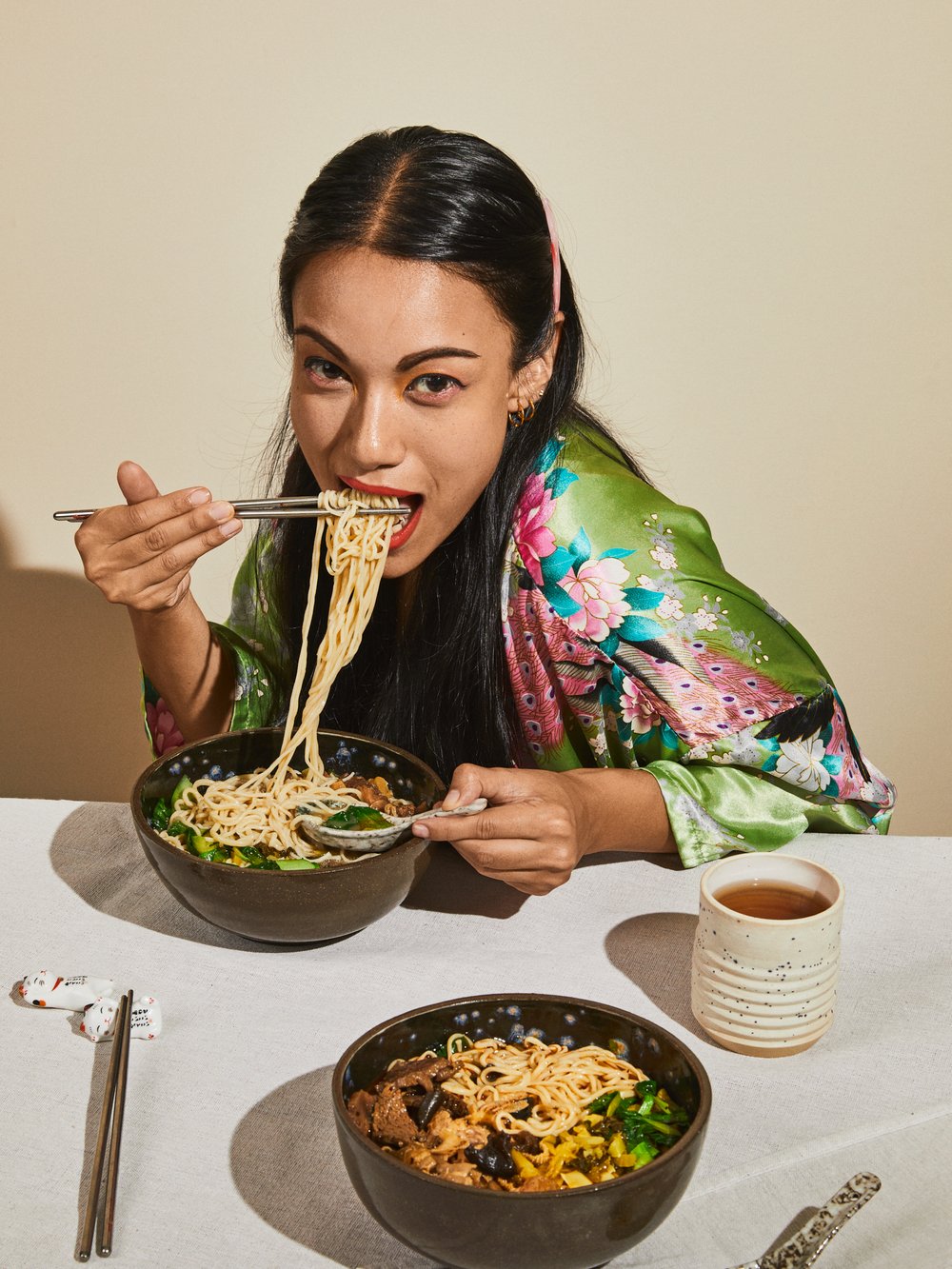 Woman eating noodles with chopsticks from a bowl at a table. She is wearing a floral robe. A cup of tea is next to another bowl.