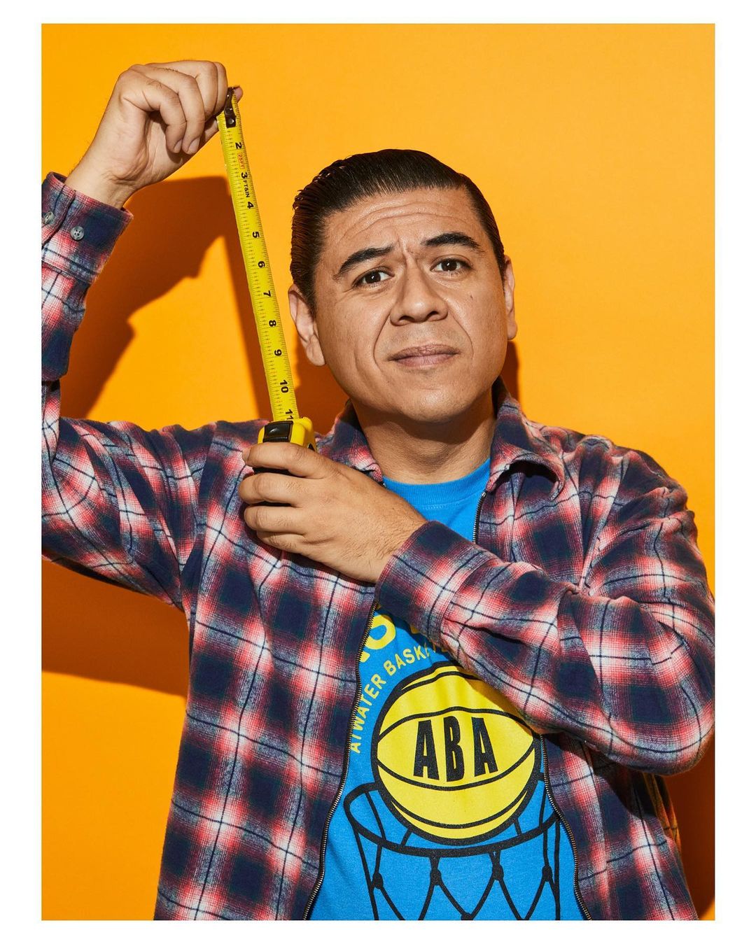 Portrait of an adult male in a blue graphic t-shirt and plaid shirt, holding a yellow measuring tape against an orange backdrop.