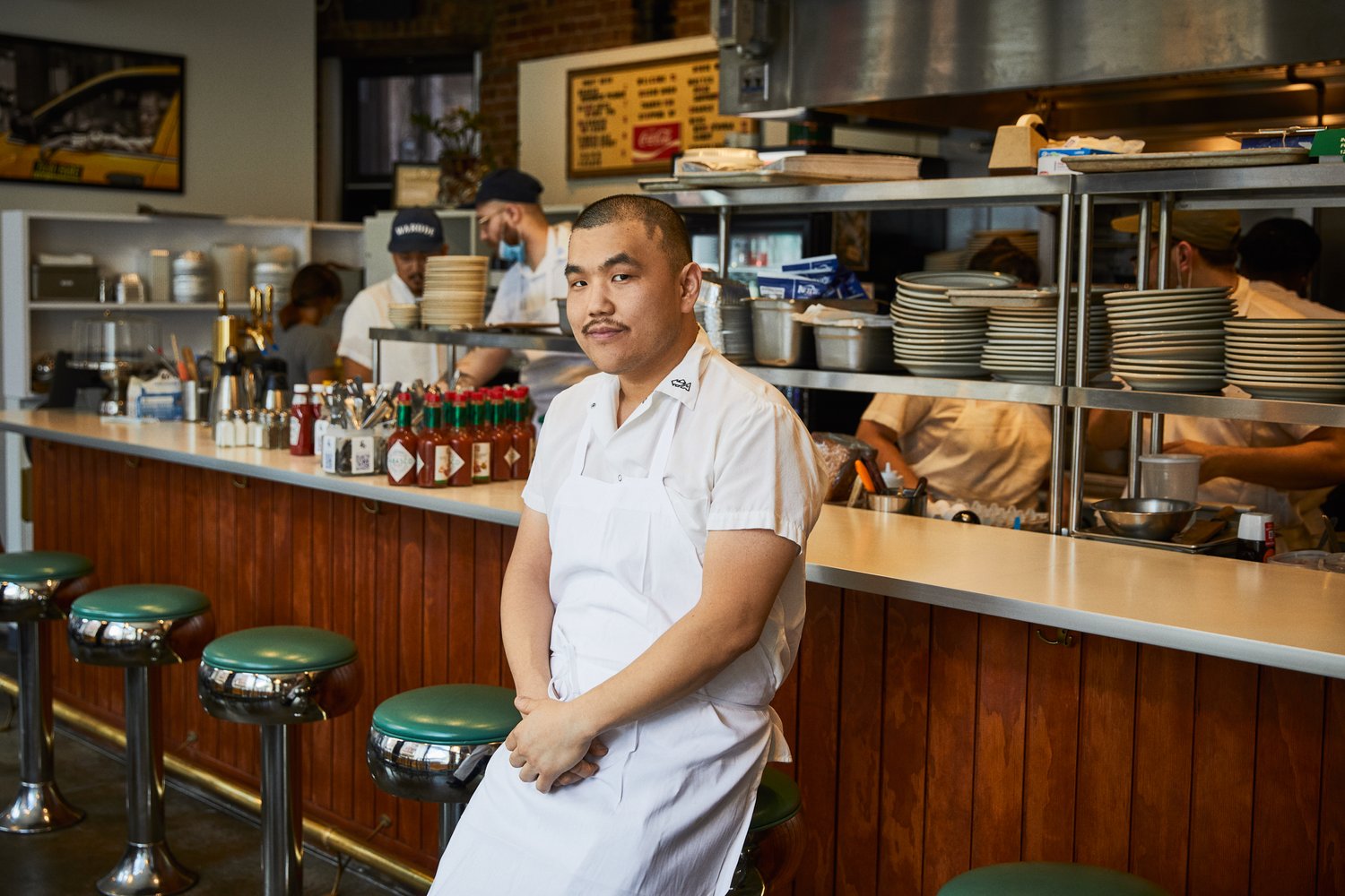 Portrait of a restaurant worker in a white apron sitting on a green stool, with other workers and dining details in the background.