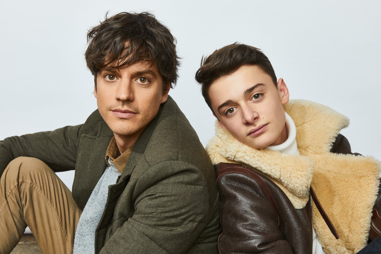 Two men pose indoors. One is wearing a green coat and the other is wearing a shearling jacket in a portrait-style medium shot.