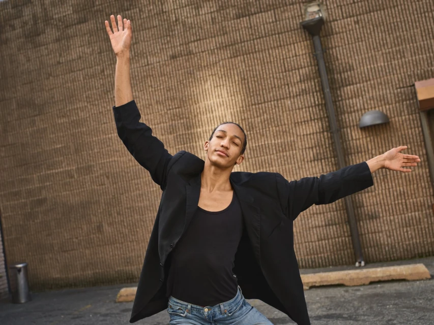 A slender person in a black blazer and jeans poses dynamically with arms raised against a brick wall backdrop outdoors.