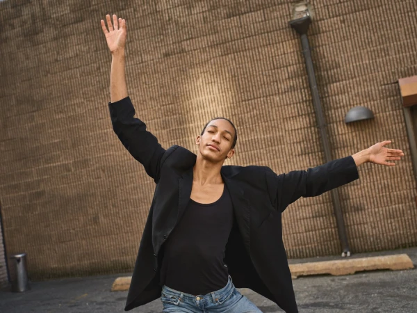 A slender person in a black blazer and jeans poses dynamically with arms raised against a brick wall backdrop outdoors.