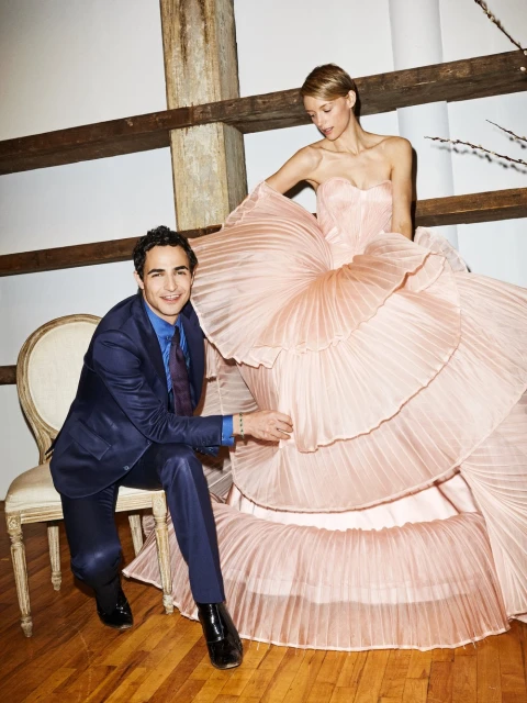 A woman in a pink ruffled gown standing next to a seated man in a blue suit, posing indoors with a neutral backdrop and wooden beam details.