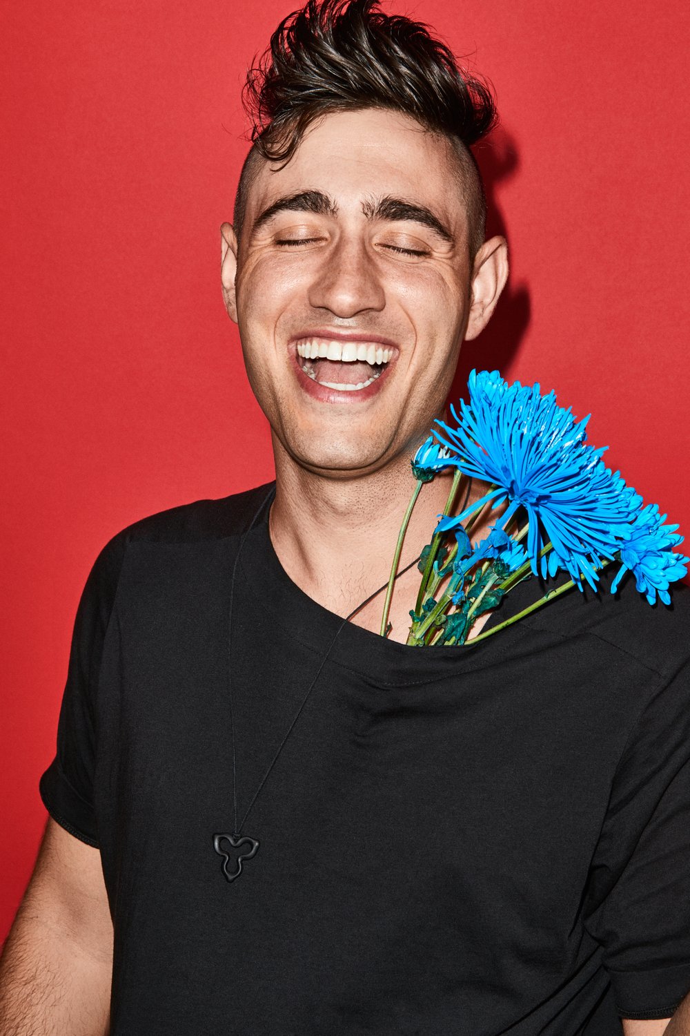 Portrait of smiling adult male with short dark hair and flowers in his shirt pocket in front of a solid red backdrop.