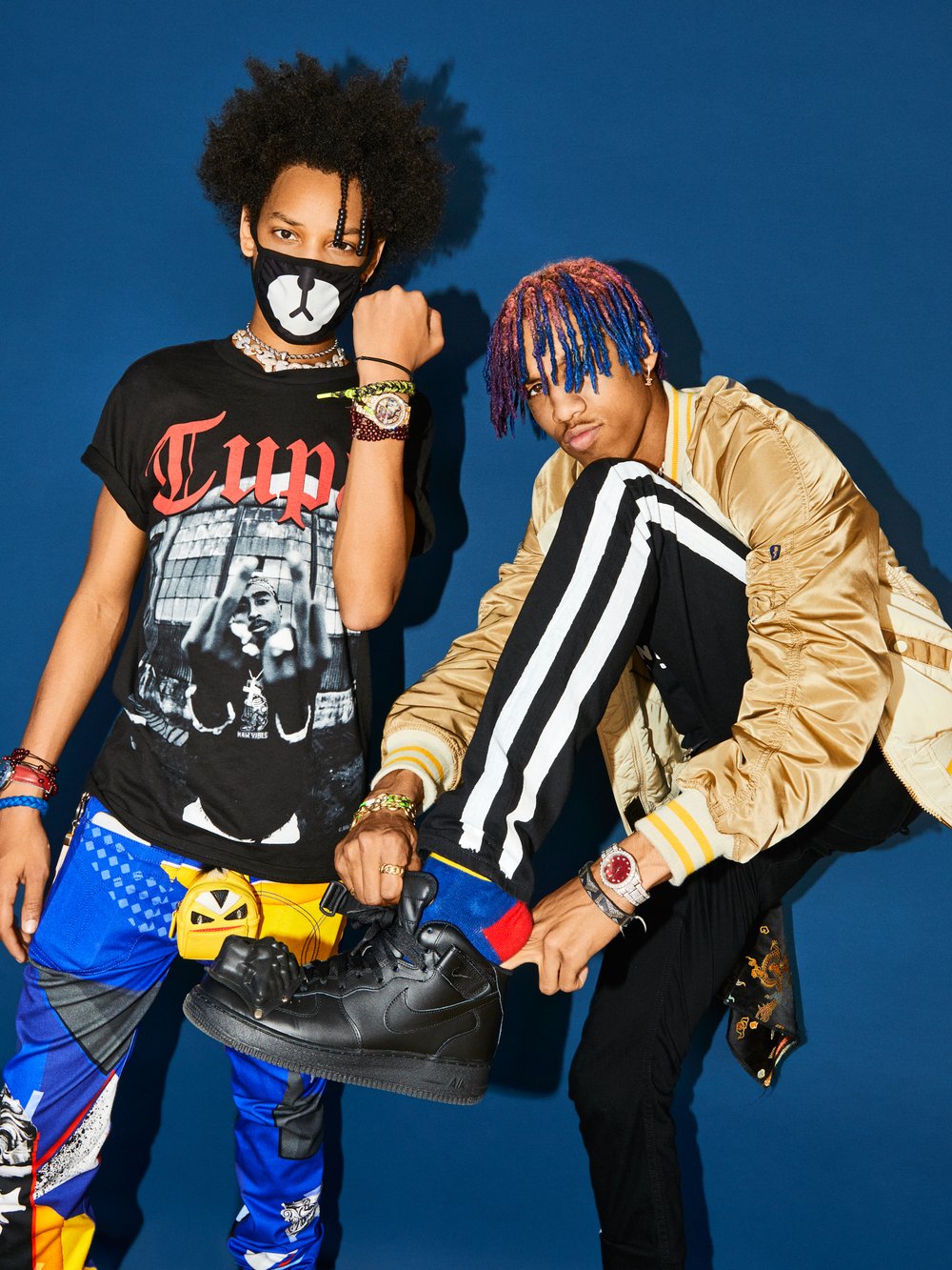 Two stylish young men pose against a blue background, one wearing a Tupac t-shirt and the other a gold jacket.