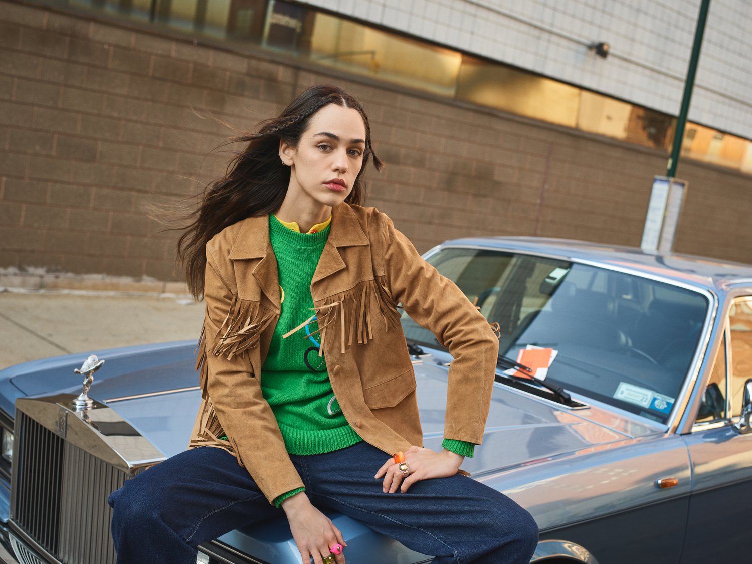 Adult woman sitting on the hood of a vintage car wearing a fringe jacket and jeans with a city street and building in the background.
