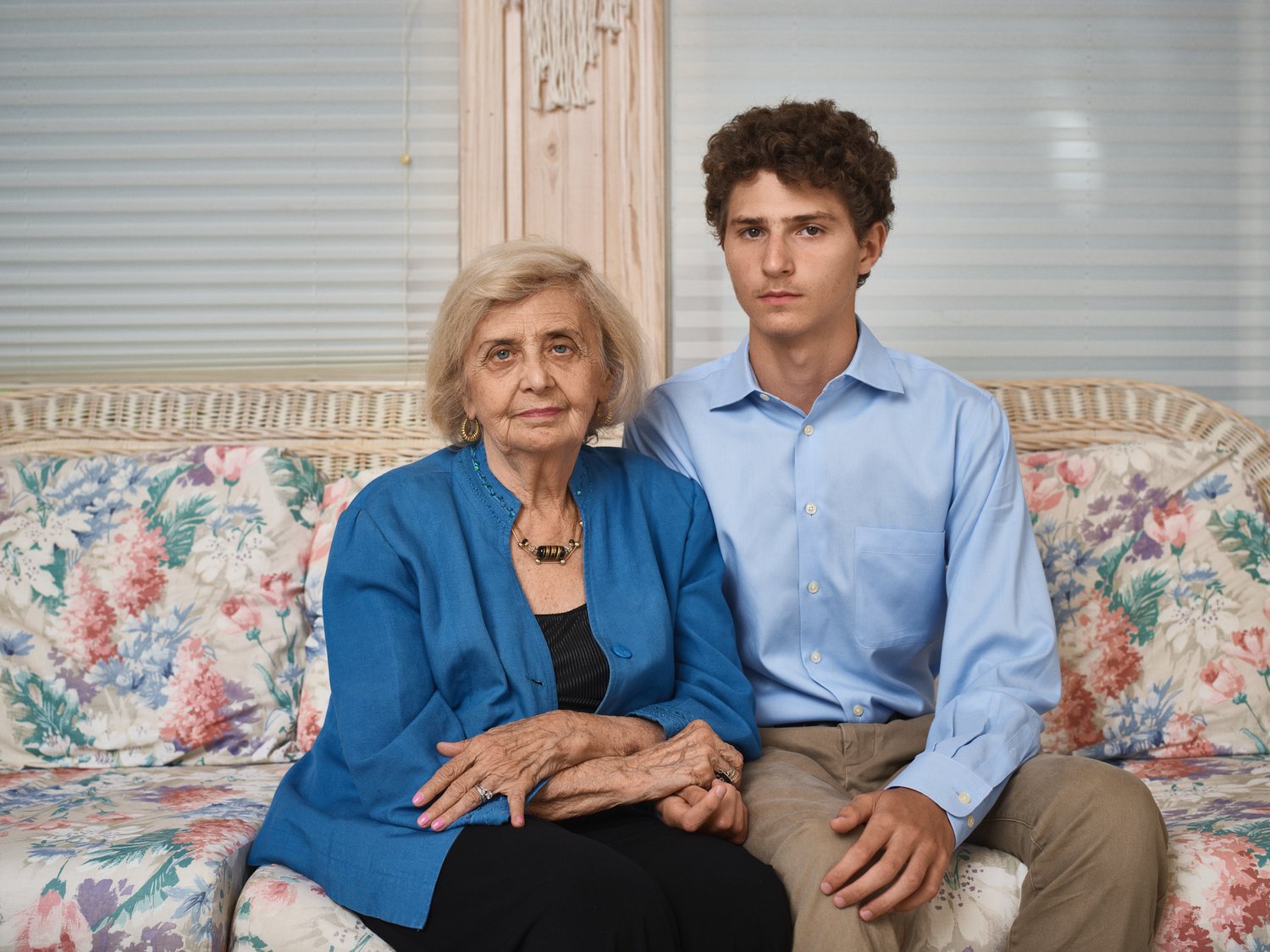 An elderly woman and a young adult man sit side-by-side on a floral sofa in a brightly lit indoor setting, looking directly at the camera.