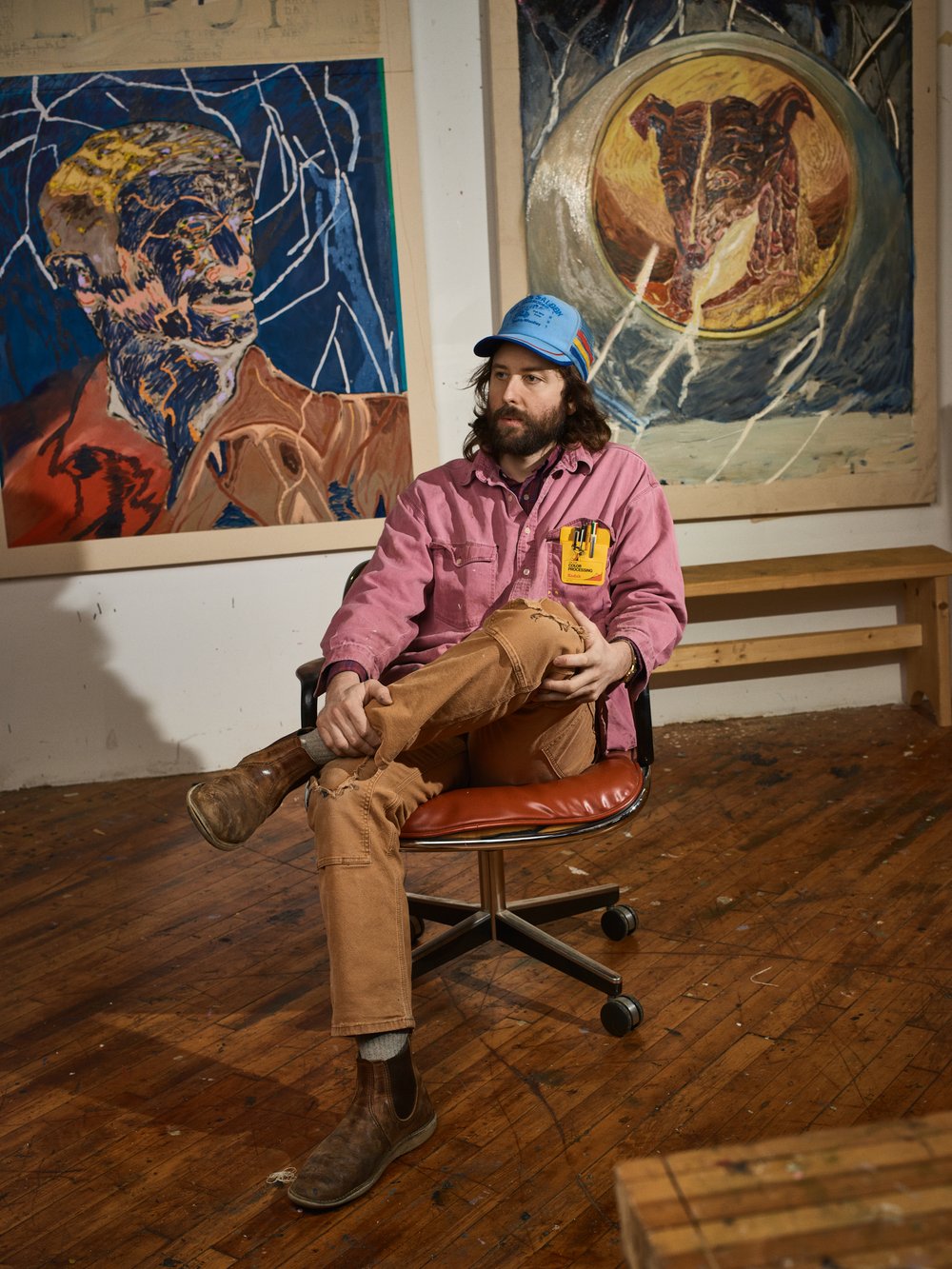 Artist sitting on a rolling chair in front of his artwork. He has a beard and is wearing a blue hat, pink jacket, and brown pants.