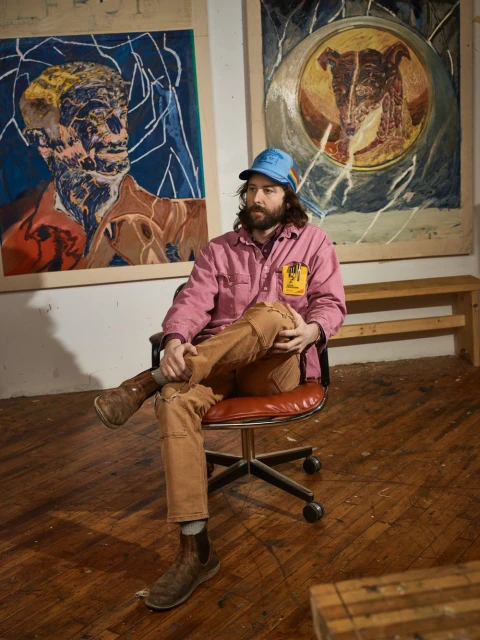 Artist sitting on a rolling chair in front of his artwork. He has a beard and is wearing a blue hat, pink jacket, and brown pants.