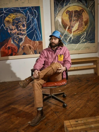 Artist sitting on a rolling chair in front of his artwork. He has a beard and is wearing a blue hat, pink jacket, and brown pants.