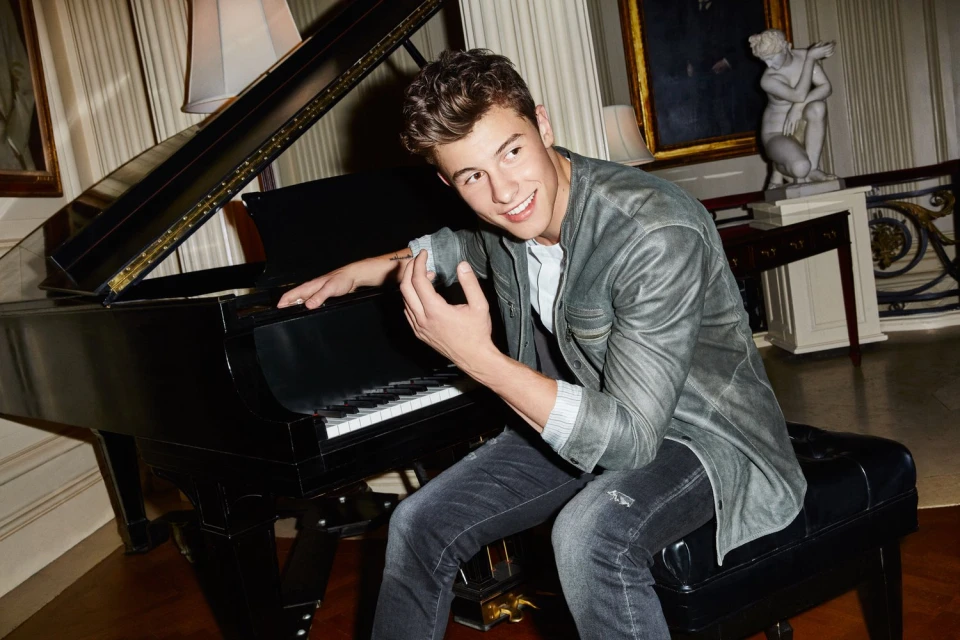 A young man with curly brown hair sits at a black grand piano in a room with white walls, a sculpture, and framed art pieces.