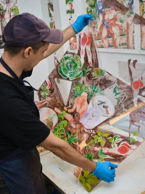 Adult male artist wearing a cap and blue gloves holds up a transparent sheet with drawings in a bright indoor studio setting.
