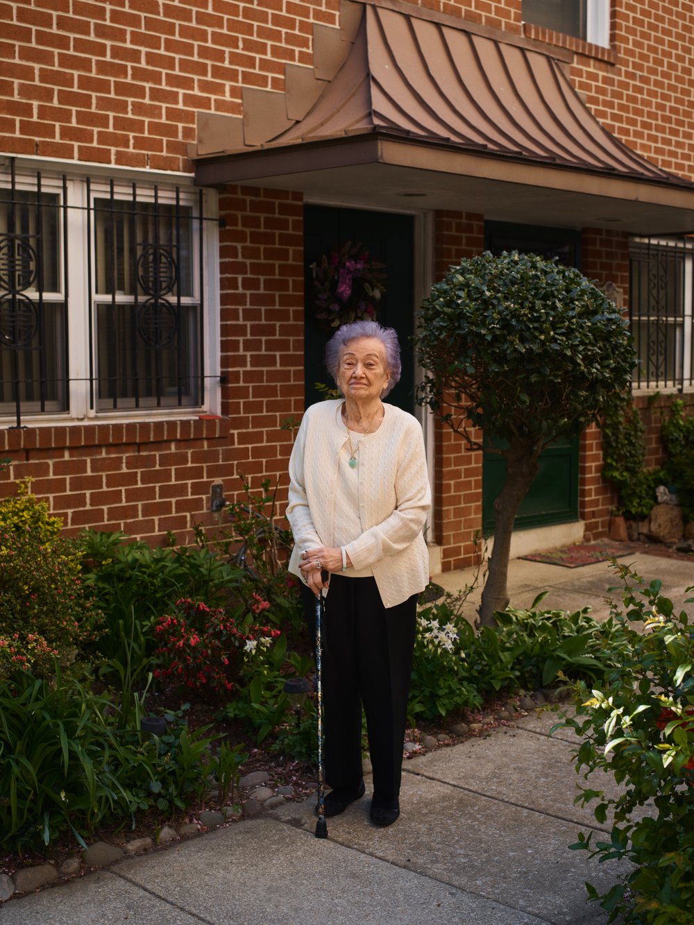 Elderly woman stands on a walkway in front of a brick building with a green bush and a cane. She is wearing a cream sweater.
