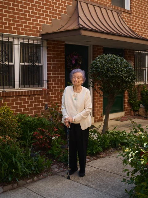Elderly woman stands on a walkway in front of a brick building with a green bush and a cane. She is wearing a cream sweater.