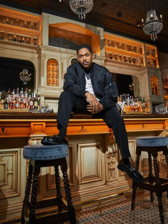 A man in a black bomber jacket and dark trousers poses seated atop a wooden bar stool inside an ornate vintage bar.