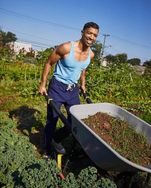 A smiling man pushes a wheelbarrow full of soil in a sunlit community garden, wearing a tank top, with greenery and houses in the background.